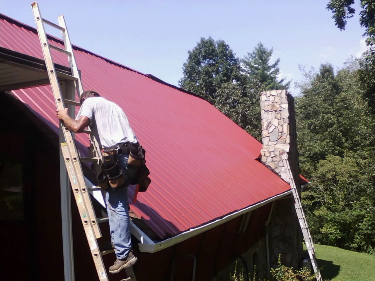 Roofer on a ladder beside a red metal roof and stone chimney — Soffit & Fascia Repair by Roofing Brothers in Hoboken